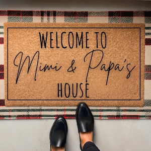 以下が含まれることがあります： 黒文字で「Welcome to Mimi & Papa's House」と書かれた茶色のウェルカムマット。マットは赤、白、緑の格子柄の敷物の上に置かれています。
