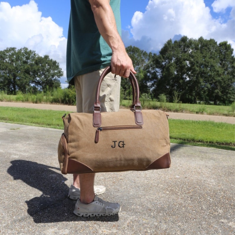 May include: A brown canvas duffel bag with brown leather accents and a zipper closure. The bag has a personalized monogram that reads "JG".