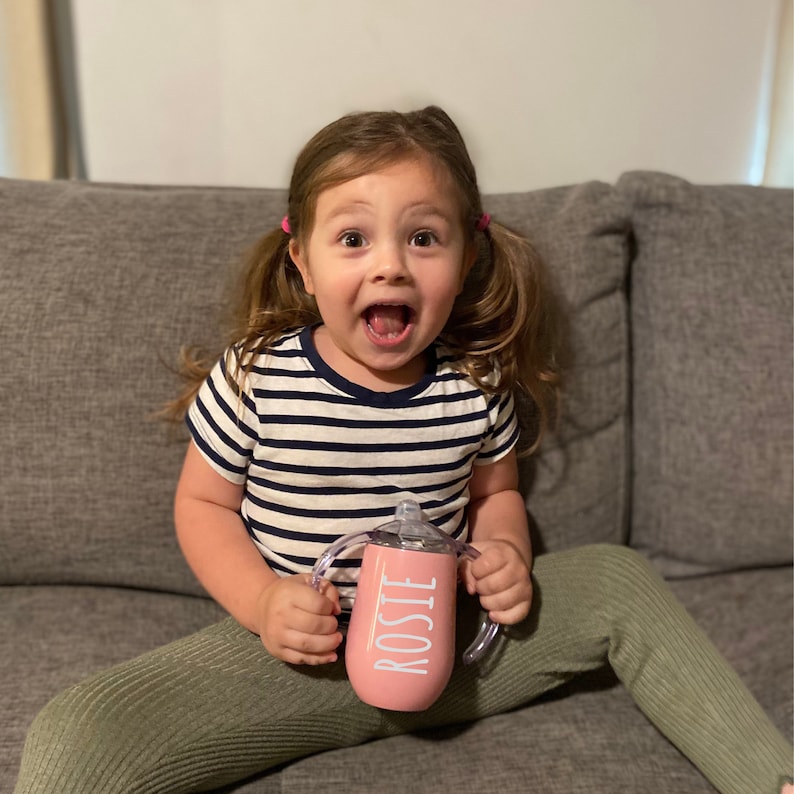 Puede incluir: Una ni&ntilde;a peque&ntilde;a con coletas est&aacute; sentada en un sof&aacute; sosteniendo una taza de beber de acero inoxidable rosa con el nombre "Rosie" impreso en ella.