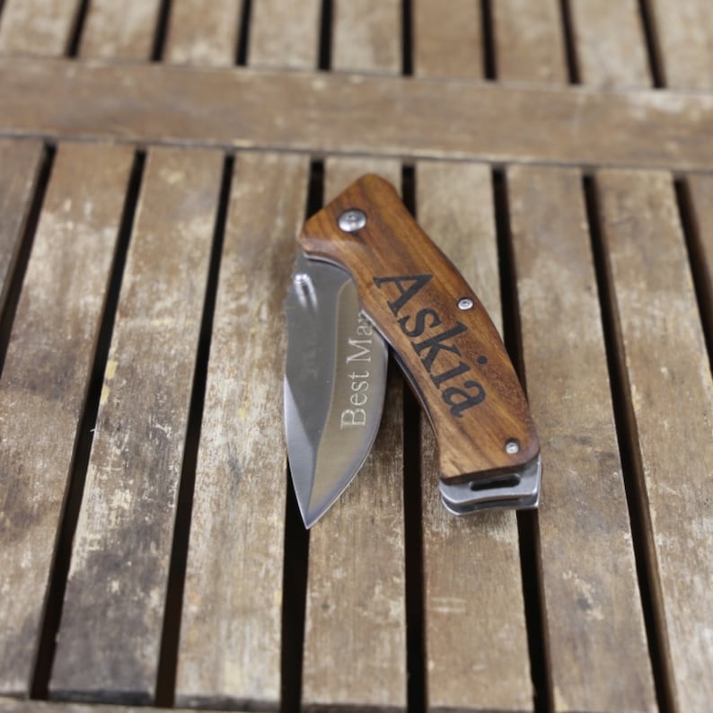 May include: A wooden handled pocket knife with a stainless steel blade. The handle is engraved with the name "Askia" and the words "Best Man".
