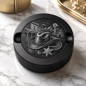 May include: A round black ashtray featuring a detailed monochromatic engraving of a cowboy hat, a coiled lasso, a sheriff star badge, and a western landscape with a windmill and wooden buildings. The design is centred on the flat top surface.