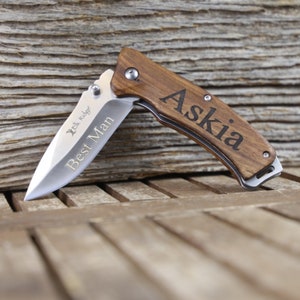 May include: A brown wooden pocket knife with a silver blade. The knife is engraved with the name "Askia" and the words "Best Man".
