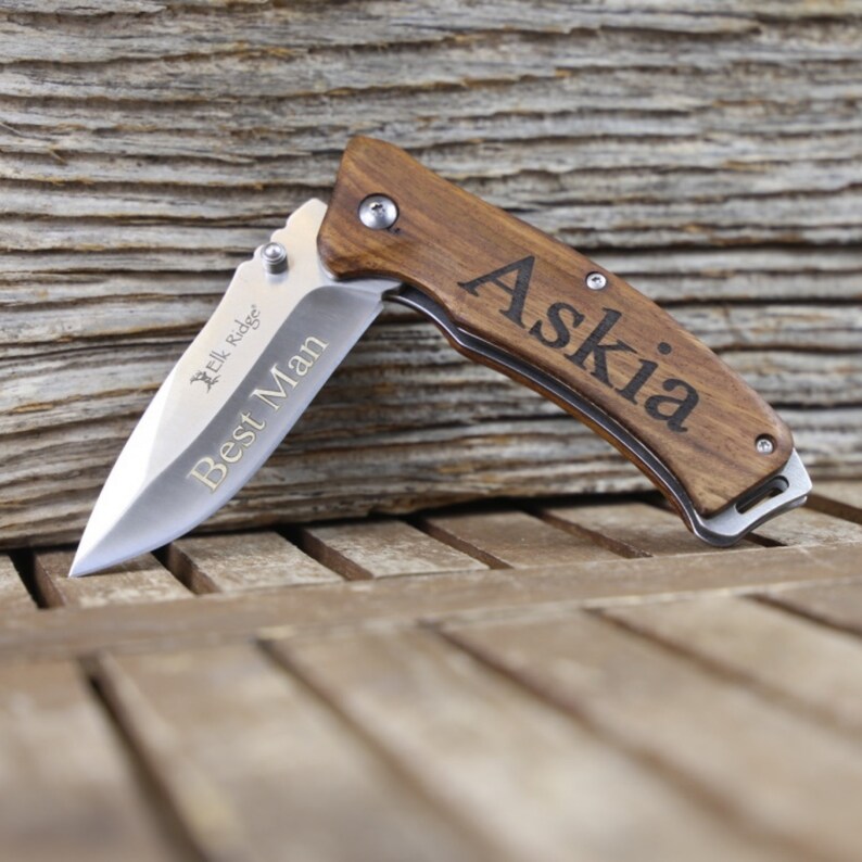 May include: A brown wooden pocket knife with a silver blade. The knife is engraved with the name "Askia" and the words "Best Man".