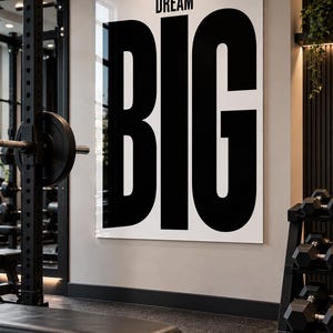 May include: A large rectangular wall art piece featuring a white background with the word DREAM in small black letters above the word BIG in very large black letters. The sign is mounted on a light gray wall inside a gym setting.