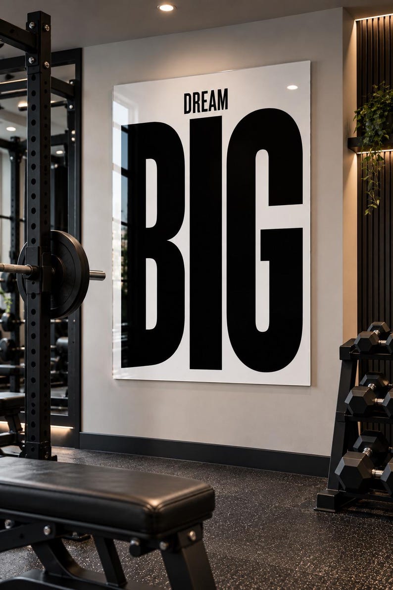 May include: A large rectangular wall art piece featuring a white background with the word DREAM in small black letters above the word BIG in very large black letters. The sign is mounted on a light gray wall inside a gym setting.