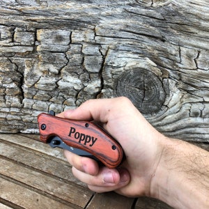 May include: A red and black pocket knife with a wood handle. The knife has the name "Poppy" engraved on the handle.