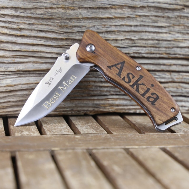 May include: A brown wooden pocket knife with a silver blade. The knife has the words "Best Man" engraved on the blade and "Askia" engraved on the handle.