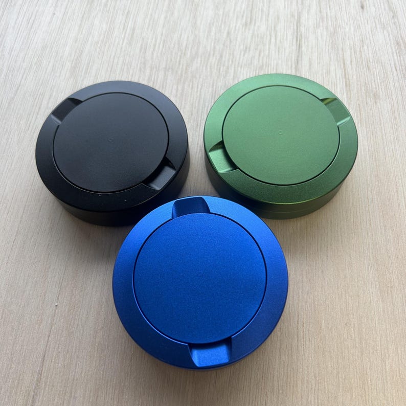 May include: Three circular containers with matte finishes in black, green, and blue. Each container features a central lid with a smooth, flat top surface and a slightly raised outer rim with two small, symmetrical notches on opposite sides.