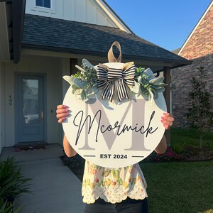 May include: A round, white wooden sign with the name "McCormick" in script and the year "EST 2024". The sign is decorated with a black and white striped bow, greenery, and a gray initial "M". A tan loop is at the top for hanging.