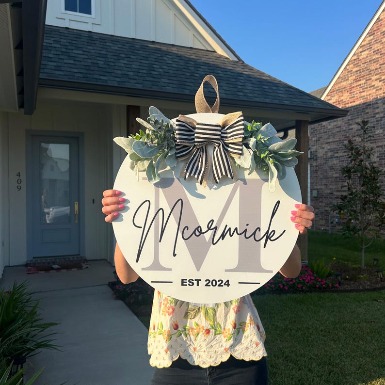 May include: A round, white wooden sign with the name "McCormick" in script and the year "EST 2024". The sign is decorated with a black and white striped bow, greenery, and a gray initial "M". A tan loop is at the top for hanging.
