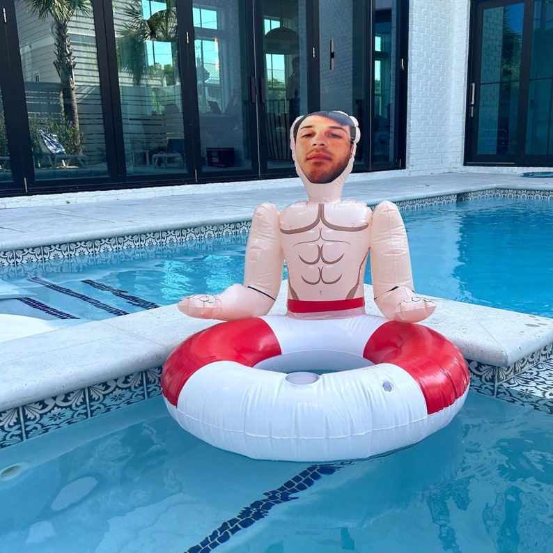 Op de afbeelding: Een witte opblaasbare zwembad drijver in de vorm van een persoon met een menselijk gezicht en een rode en witte reddingsboei om de taille. De drijver bevindt zich in een zwembad.