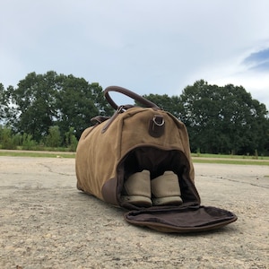 May include: A brown canvas duffel bag with leather accents and a zippered opening. The bag is open and two pairs of white sneakers are visible inside.