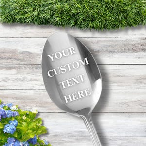May include: A silver spoon with a personalized message that reads "YOUR CUSTOM TEXT HERE". The spoon is on a white wooden surface with green foliage in the background.
