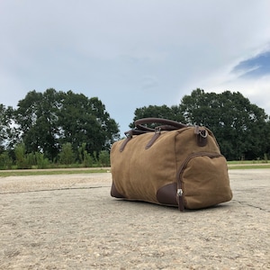 May include: A brown canvas duffel bag with leather accents and a zipper closure. The bag is sitting on a concrete surface.
