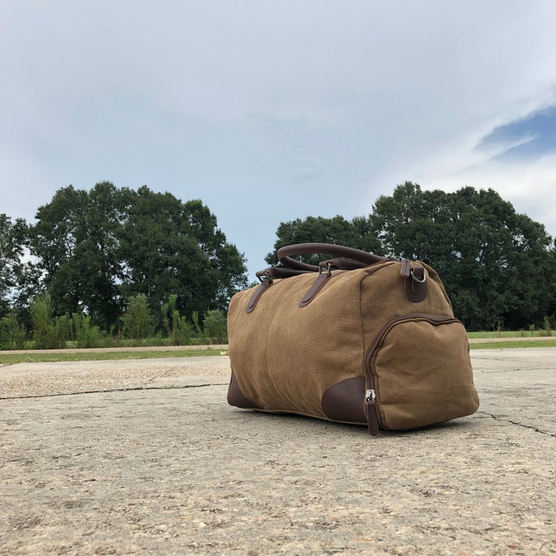 May include: A brown canvas duffel bag with leather accents and a zipper closure. The bag is sitting on a concrete surface.