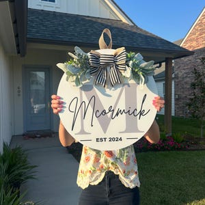 May include: A round, white wooden sign with the name "McCormick" in cursive script and a large letter "M" in gray. The sign is decorated with greenery, a striped bow, and the text "EST 2024".