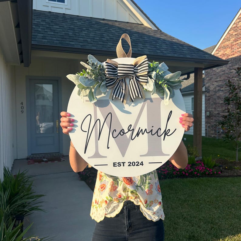 May include: A round, white wooden sign with the name "McCormick" in cursive script and a large letter "M" in gray. The sign is decorated with greenery, a striped bow, and the text "EST 2024".