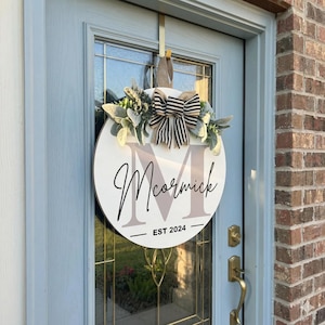 May include: A round, white wooden door wreath with the name "McCormick" in black script and the initial "M" in a light brown color. The wreath is decorated with greenery, a striped bow, and the text "EST 2024". It hangs on a light blue door.