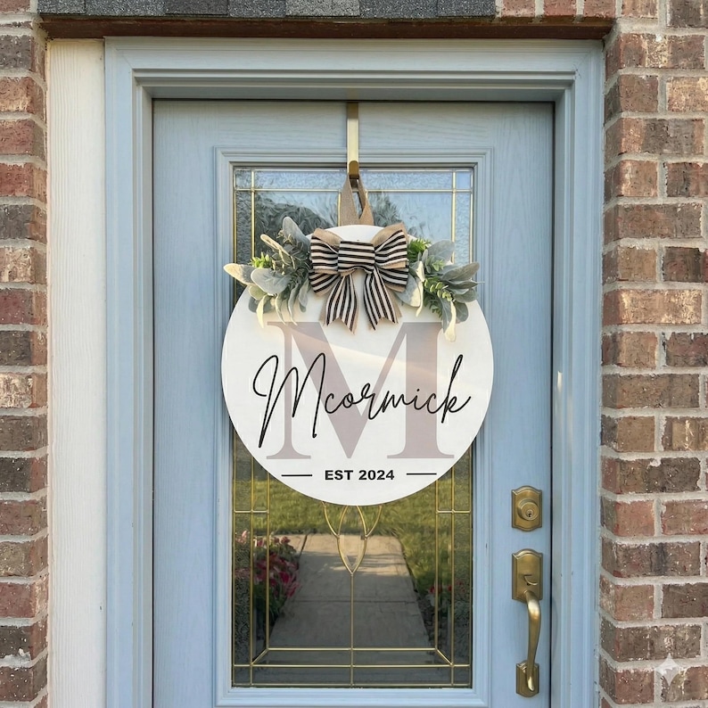 May include: A round, white wooden door sign with the name "McCormick" in elegant script. The sign features a large letter "M" and the text "EST 2024". It is adorned with a striped bow and greenery.