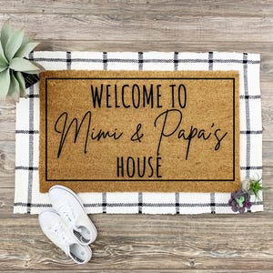 以下が含まれることがあります： 黒文字で「Welcome to Mimi & Papa's House」と書かれた茶色のココナッツ製の玄関マット。マットは白黒の市松模様の敷物の上に置かれています。