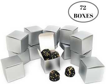 Puede incluir: 72 cajas de regalo plateadas, cada una con forma cuadrada y un interior blanco. Las cajas están apiladas en forma de pirámide, con algunas cajas abiertas y llenas de pequeños chocolates negros y dorados.