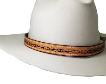 May include: A white cowboy hat with a decorative leather hatband. The hatband is light brown with a detailed black pattern and white stitching. The hat has a wide brim and a rounded crown.