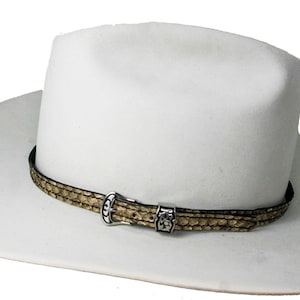 Puede incluir: Un sombrero de vaquero blanco con una banda de cuero marrón y una hebilla plateada. La banda tiene un patrón de piel de serpiente.
