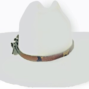 Op de afbeelding: Een witte cowboyhoed met een decoratieve band. De band heeft een bruin, beige en zwart patroon. De hoed heeft een klassieke, afgeronde kroon en een brede rand.