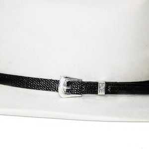 May include: A white cowboy hat with a black textured band and silver-colored buckle. The hat band features a decorative buckle and a small silver-colored accent piece. The hat is a classic Western style.