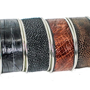 May include: A set of eight leather cuff bracelets in various colors and patterns, including brown, black, and orange. Each bracelet has a silver metal band around the edge.