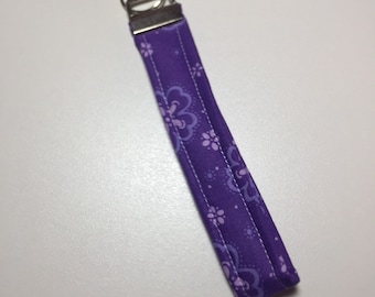 May include: A purple fabric wristlet key fob with a floral pattern in shades of purple and white. The key fob has a silver metal clasp and a split ring for attaching keys.