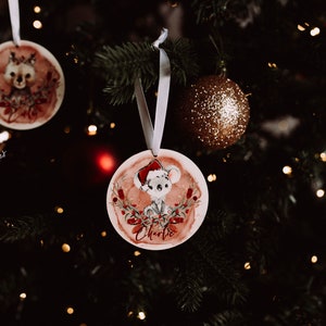 May include: Two round Christmas ornaments with watercolor illustrations of animals. The first ornament features a deer with antlers and the text "Charlie". The second ornament features a koala wearing a Santa hat and the text "Charlie".