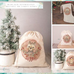 May include: A set of three personalized Christmas gift bags with a drawstring closure. The bags feature a watercolor illustration of an Australian animal with a wreath around its head. The bags are beige and have the names "Maggie Mae", "The Corden Family", and "Maggie" printed on them.