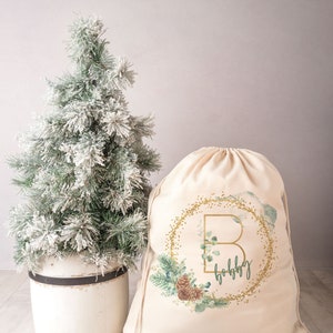 May include: A white canvas drawstring bag with a gold glitter and green foliage design. The bag features a gold letter "B" in a wreath with the word "ho ho ho" written below it.