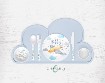 May include: A blue silicone placemat shaped like a cloud with a white airplane design on a blue plate. The plate has the name "Bobby" written on it. The set includes a spoon, fork, knife, and two small bowls.