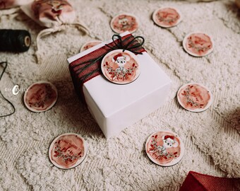 May include: A white gift box with a red ribbon and a watercolor-style Christmas ornament featuring a mouse wearing a Santa hat. The ornament has the name "Charlie" written on it. The box is surrounded by other similar ornaments with various designs.