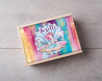 Può includere: Una scatola di legno con un design di sirena colorato. La scatola ha uno sfondo arcobaleno con glitter dorati e una sirena seduta in una conchiglia. La sirena tiene un piccolo cavalluccio marino e c'è un delfino che nuota sopra di lei. La scatola è etichettata "Josette".