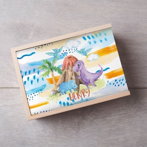 May include: A wooden box with a colorful design featuring dinosaurs, a volcano, and palm trees. The box has the name "HENRY" written on the front.