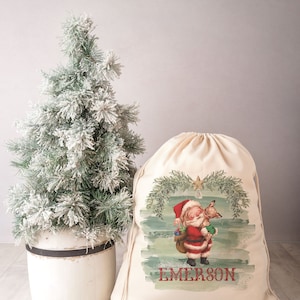 Könnte beinhalten: Ein weißer Stoff-Weihnachtsmann-Sack mit Kordelzug. Der Sack zeigt eine Vintage-Illustration von Weihnachtsmann, der ein Rentier hält, vor einem grün-blauen Aquarellhintergrund. Der Name "EMERSON" ist unten auf dem Sack in Rot gedruckt.
