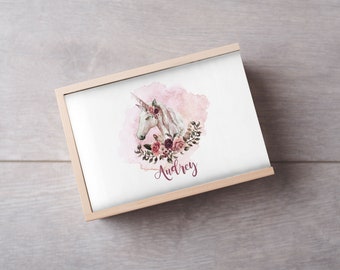 Può includere: Una scatola di legno con un coperchio bianco che presenta un'illustrazione ad acquerello di un unicorno con una corona di fiori. L'unicorno è circondato da acquerello rosa e il nome "Audrey" è scritto in corsivo sotto l'immagine.