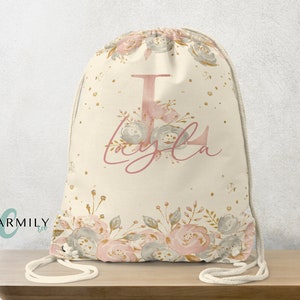 Puede incluir: Una bolsa de cordón blanco con un diseño floral y purpurina dorada. La bolsa tiene una gran letra "L" rosa y el nombre "Layla" escrito en cursiva rosa.