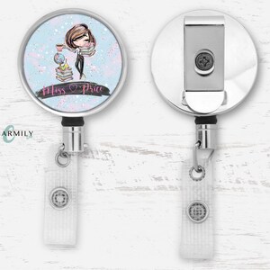 May include: A silver retractable badge reel with a round, blue and white design featuring a cartoon woman holding books. The design also includes the text "Miss Price" with a pink heart.