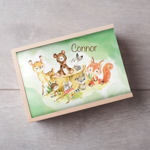 Pode incluir: Uma caixa de madeira com fundo verde e uma ilustração de aquarela de um veado, um urso, um esquilo e outros animais da floresta. A caixa tem o nome "Connor" escrito nela.