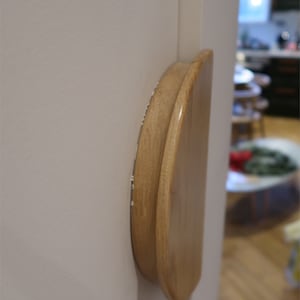 May include: A wooden door handle with a curved, half-moon design. The handle is made of light-colored wood and is mounted on a white door. The handle's smooth surface contrasts with the textured wall. The door is slightly ajar, revealing a glimpse of the interior.