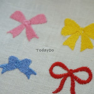 May include: Four embroidered bow appliques in pink, yellow, blue, and red. The bows are stitched onto a white fabric. The text "TodayDo" is printed on the fabric.