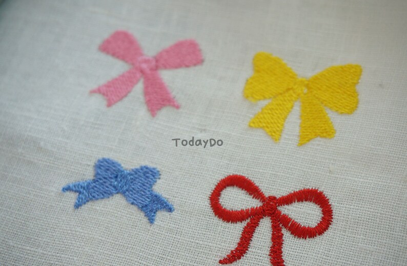 May include: Four embroidered bow appliques in pink, yellow, blue, and red. The bows are stitched onto a white fabric. The text "TodayDo" is printed on the fabric.
