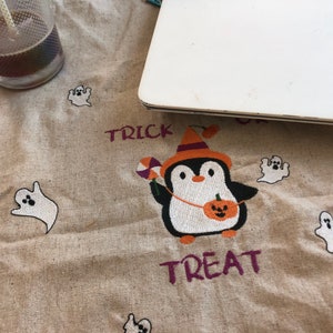 May include: A beige Halloween-themed fabric with embroidered elements. A penguin in a witch hat holds a pumpkin and candy. The words "Trick" and "Treat" are embroidered in purple. White ghost designs are scattered across the fabric.
