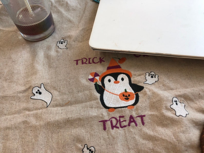 May include: A beige Halloween-themed fabric with embroidered elements. A penguin in a witch hat holds a pumpkin and candy. The words "Trick" and "Treat" are embroidered in purple. White ghost designs are scattered across the fabric.