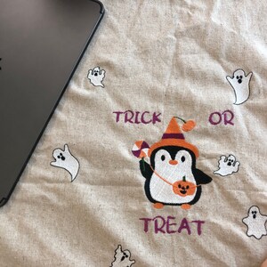 May include: A beige Halloween-themed fabric with embroidered elements. Features a penguin in a witch hat holding candy and a pumpkin, surrounded by white ghost figures. The words "TRICK OR TREAT" are embroidered in purple.