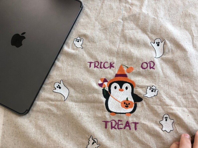 May include: A beige Halloween-themed fabric with embroidered elements. Features a penguin in a witch hat holding candy and a pumpkin, surrounded by white ghost figures. The words "TRICK OR TREAT" are embroidered in purple.